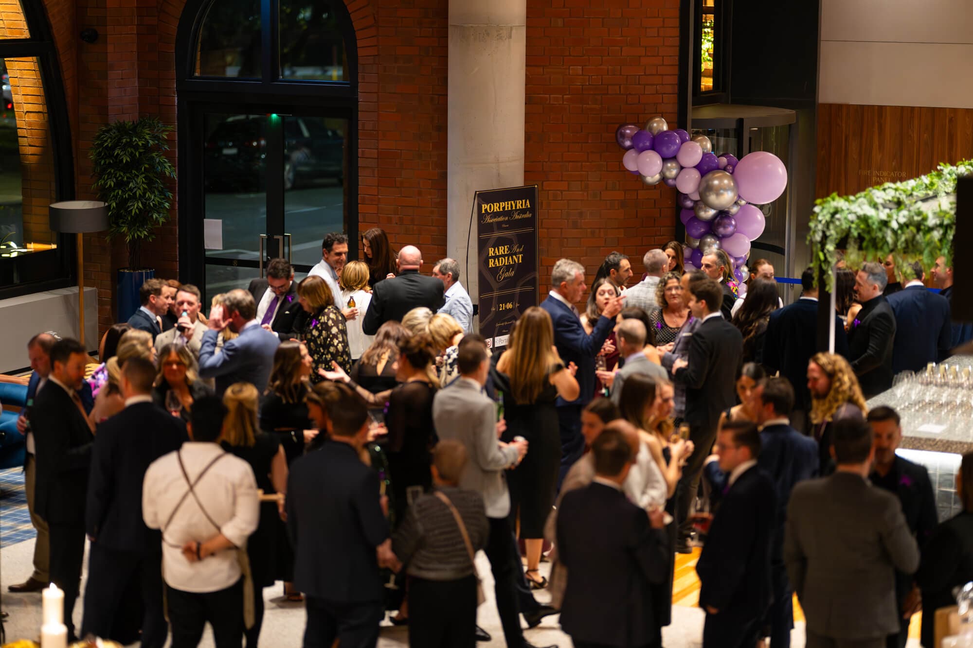 A full room of guests mingling at the 2025 gala, purple balloon garland in the background